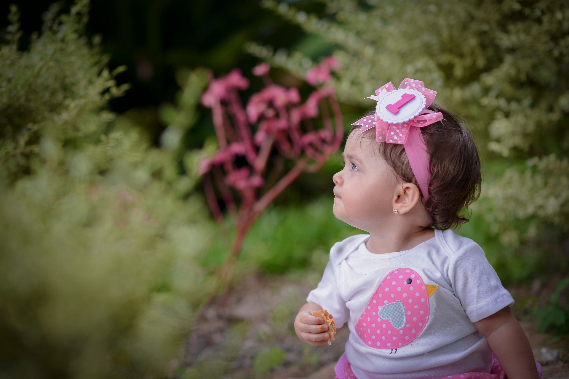 Cute baby sitting on meadow looking up as she holds candy in her hand.